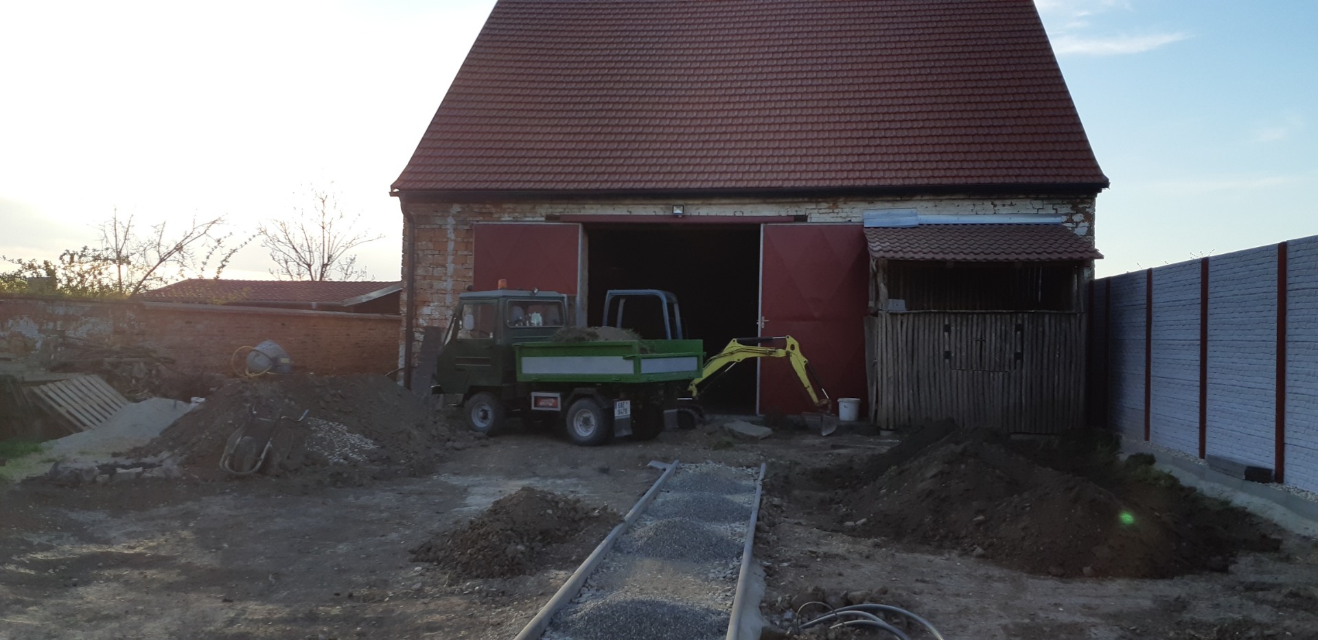 Multicar s bagrem při zemních pracích u stodoly na venkově